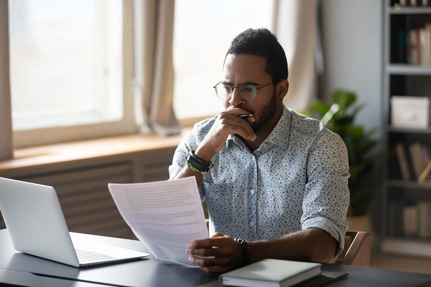 Thoughtful young man in glasses holding paper document, reading banking debt notification.
